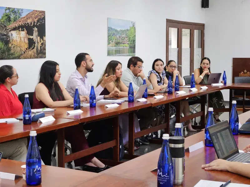 Un grupo de profesionales de la salud y residentes de Medicina Familiar participan en una mesa de trabajo durante el Gran Simposio de Medicina Familiar en la Javeriana Cali. Sentados alrededor de una mesa en forma de “U”, conversan y toman apuntes mientras trabajan colaborativamente. Sobre la mesa hay botellas de agua, portátiles y cuadernos. Al fondo se observan fotografías de comunidades rurales, que acompañan este espacio académico de reflexión y actualización clínica.
